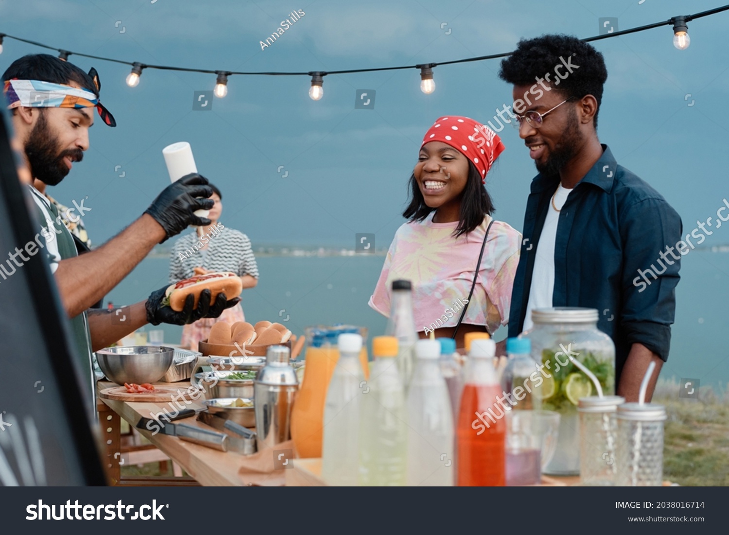 stock-photo-african-young-couple-watching-for-the-process-of-preparing-hot-dogs-they-having-lunch-on-the-beach-2038016714