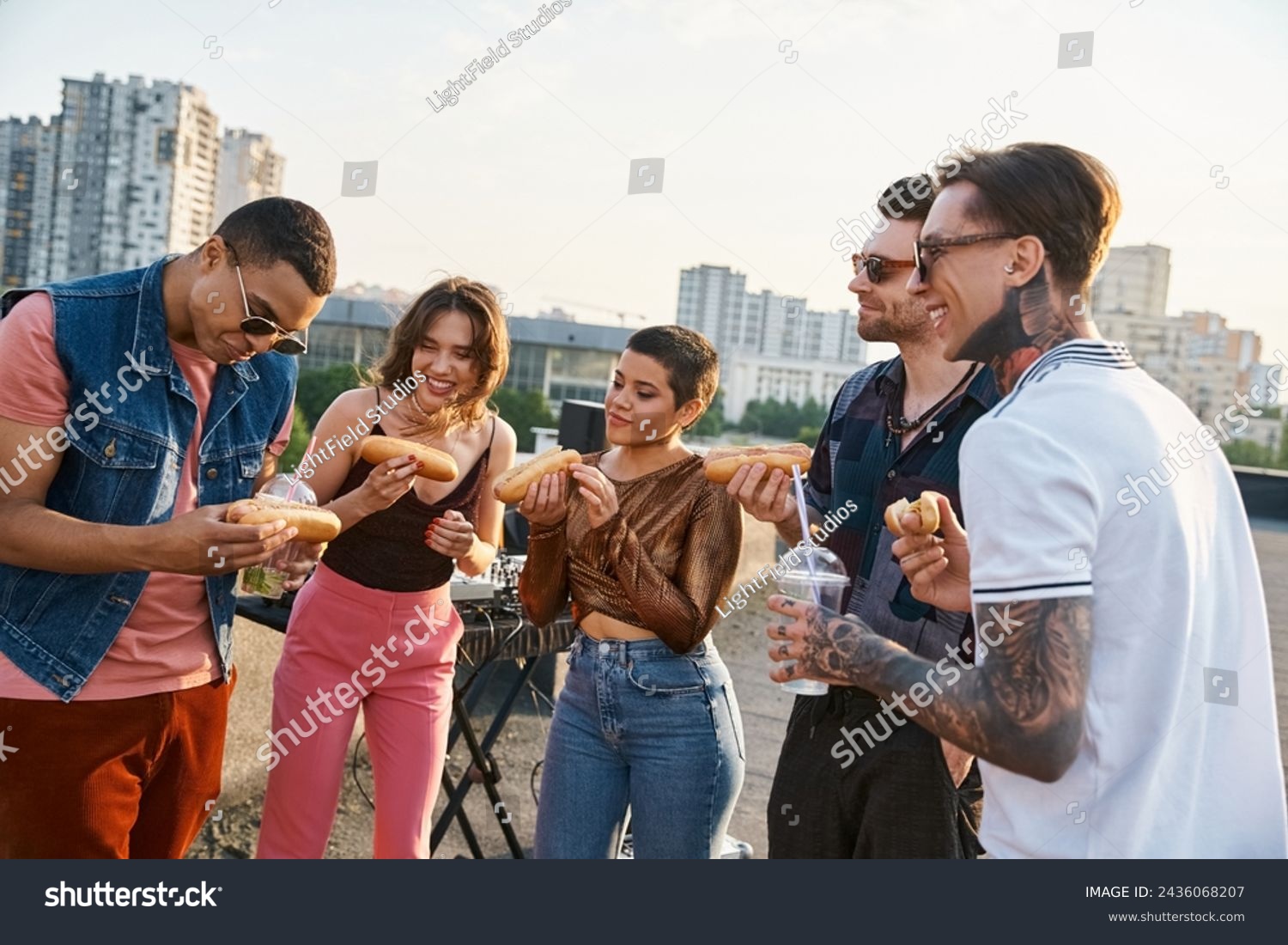 stock-photo-good-looking-jolly-multicultural-friends-in-urban-outfits-eating-tasty-hot-dogs-at-rooftop-party-2436068207